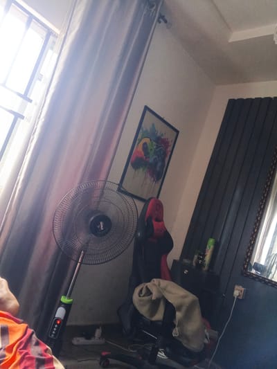 The media file depicts an indoor scene, likely a bedroom or living area, in Naze, Nigeria. Sunlight streams in from a large window on the left, partially obscured by light-colored curtains. Dominating the foreground is a black oscillating fan with a silver stand. The fan has a circular grill and a central black circular component with a logo. At the base of the fan's stand, a green and black control panel is visible, showing a red digital display.
To the right of the fan, a black and red gaming chair is positioned, draped with a beige fabric, possibly a blanket or throw. Behind the chair, a framed piece of art hangs on the wall, featuring a colorful depiction of a video game controller with abstract splashes of paint. Further to the right, a dark cabinet or dresser is visible, with a green spray can and other items on its surface. A large, dark-colored vertical blind covers a portion of the wall on the far right, and a mirror with an ornate frame is also present.
The overall lighting suggests it is daytime, with the bright sunlight indicating clear weather. The presence of a gaming chair and artwork related to video games hints at the occupant's interests. The scene is somewhat cluttered with electrical cords and a power strip visible near the floor. The angle of the shot and the presence of a person's leg, covered in a striped garment, in the lower left corner suggest the photo was taken by someone within the room. No explicit emotions are conveyed beyond a sense of everyday domesticity. There is a visible logo on the fan which is partially obscured but appears to read "POW".