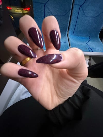 A close-up shows a person's hand with long, almond-shaped nails painted a deep burgundy color with subtle glitter flecks. The hand has a gold ring on one finger and a silver ring on another. The background includes a blue and gray surface, a glimpse of a car, and a dark fabric sleeve. The focus is on the hand and the glossy, jewel-toned nails.