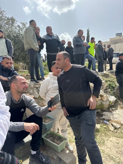 The scene depicts an outdoor gathering of approximately 15-20 adult males in the city of Dura, Palestinian Territory, under a partially cloudy sky. In the mid-right foreground, a man in a black long-sleeved shirt with an Under Armour logo and dark jeans stands, facing left with his left hand in his pocket. To his immediate left and slightly behind, a younger man in a white hooded sweatshirt and light-colored pants is partially visible, facing forward. In the lower left, a man in a grey and black long-sleeved shirt with an Adidas logo and black pants is crouching, holding a black smartphone horizontally in his right hand, pointed towards the standing man.
Behind these individuals, numerous other men are dispersed across uneven, rocky terrain. They are casually dressed in a variety of jackets, hoodies, and jeans. Notable figures include a man in a light green puffer jacket interacting with another in a grey and black hoodie in the mid-left background, and a man in a bright neon yellow shirt further back on the right. The ground consists of dirt patches, scattered stones, and sparse green vegetation. Two light green rectangular planters containing soil and minimal plant growth are positioned on the ground between the crouching man and the standing men. A leafy green tree or bush is visible on the far left. In the far background, light-colored buildings, possibly residential structures, and a slender evergreen tree are present under the open sky.