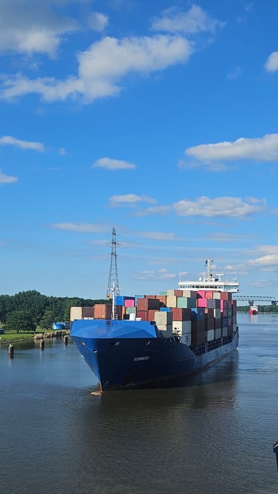 This image depicts the container ship ELBWATER navigating a waterway in Brunsbüttel, Germany, under a bright blue sky dotted with white clouds. The ship, laden with stacked shipping containers of various colors, is positioned centrally in the frame, moving forward. Its blue hull is visible, with the name "ELBWATER" clearly marked on its side.

To the left of the ship, a tall electrical pylon stands prominently against the sky, with a bridge structure visible in the background to the right. A second, smaller red ship is seen in the distance, further down the waterway. The water is a dark, rippling surface, reflecting the sky. Along the banks, lush green trees form a dense line, with some dark cylindrical buoys or markers visible in the water near the left bank. A few scattered buildings, including what appear to be blue storage containers, are visible on the far bank among the trees. The overall scene suggests a sunny day with active maritime traffic.