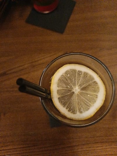An overhead shot captures a glass containing a beverage and a lemon slice, resting on a wooden table. The glass, positioned in the lower-center of the frame, holds a dark liquid, possibly iced tea or a cocktail, with a round slice of lemon floating on the surface. The lemon slice is bright yellow with a white rind and clearly shows its internal segments. To the left of the lemon, two dark, slender straws are partially submerged in the drink.
The table surface, which takes up most of the background, is made of wood with visible grain patterns and a warm, reddish-brown hue. In the upper-left corner of the image, out of focus, another object is visible: a portion of what appears to be a glass with a red-colored drink, sitting on a dark, square coaster. The overall lighting is dim, suggesting an indoor setting like a bar or restaurant, possibly in the evening. The image has a slightly grainy texture, particularly noticeable in the darker areas of the wooden table. There is no visible text in the image.