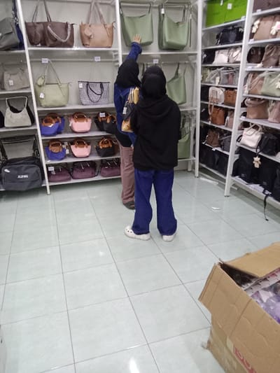 The image depicts an indoor retail environment, specifically a store featuring handbags, located in the city of Kauman, Indonesia. Two individuals are viewed from the rear, standing on a light-colored tiled floor. The person on the left wears a dark head covering, a dark blue upper garment, and light brown pants, extending their right arm towards a light green handbag displayed on an upper white metal shelf. The person on the right wears a black hooded sweatshirt, blue pants, and white clogs, holding multiple small, light-colored items in their hands. White modular shelving units line the store, showcasing a dense arrangement of handbags in various styles, sizes, and colors, including shades of green, brown, pink, blue, black, and cream. A black duffel-style bag labeled "JILIPING" is visible on a lower shelf in the bottom-left of the frame. A partially open brown cardboard box containing merchandise is situated on the floor in the bottom-right.