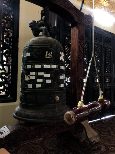 The image shows a large, dark bronze bell hanging from a wooden beam inside what appears to be a temple or pagoda in Nam Ban, Vietnam. The bell is ornate, with intricate carvings and several white labels adhered to its surface. To the right of the bell is a wooden mallet, suspended by ropes, which is used to strike the bell. The background features dark, intricately carved wooden screens, and the floor is covered in a patterned tile. The lighting suggests an indoor environment, possibly illuminated by natural light filtering through the screens and artificial lighting from above. A small stack of papers or a booklet is visible on a carved wooden surface in the lower left corner.
