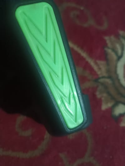 A close-up shows a black object with a bright green, rectangular insert. The green insert features three downward-pointing chevron patterns. The object appears to be made of molded plastic or rubber. A red and brown patterned rug is visible in the background, slightly blurred. The lighting highlights the texture and contours of the object.