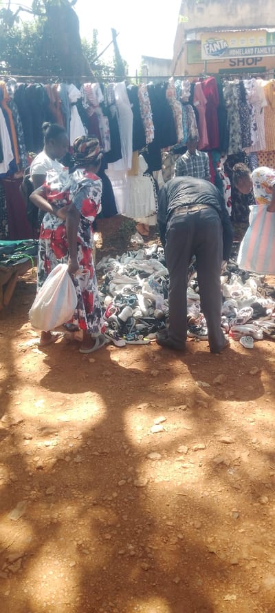 The image portrays an outdoor market scene in Shirungu-Mugai ward, Kenya, under bright, natural light with dappled shadows cast upon reddish-brown dirt and gravel ground. In the midground, a substantial, unorganized pile of various used footwear, including sneakers and sandals, is spread directly on the ground.
At least five individuals are present. Three are actively engaged with the shoe pile: a woman on the left, wearing a patterned dress and a headscarf, holds a white bag and looks down at the shoes. Beside her, a man with his back to the camera, dressed in a dark shirt, dark trousers, and a brown belt, is bent over, examining the footwear. To his right, a partially visible woman also bends over the pile, holding a striped bag. In the background, a man in a plaid shirt is partially obscured behind the shoe display.
Above the shoe pile, a metal rack extends horizontally, laden with numerous hanging garments, including dresses and tops of assorted colors and patterns. In the upper right background, a light-colored building is visible, featuring a prominent orange and white "FANTA" sign, along with a "HOMELAND FAMILY SHOP" sign below it. Green tree foliage is visible in the upper left and central background.