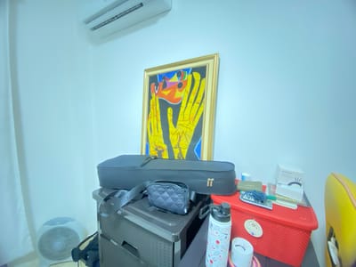 The image depicts an indoor scene with a cluttered surface. Above the objects is a painting with yellow hands on a black background and a gold frame, hanging on a light blue wall. Above the painting is a white air conditioner unit. Below the painting on a black drawer unit sits a black violin case with a small black purse resting on it. To the right of the violin case is a red plastic basket filled with various items, including a "Pandora" branded box, a teal-colored cord, and a white bottle with a black lid. A small white fan is in the left side of the image next to the drawer unit. The environment suggests a residential setting. The city of La Aurora, Ecuador, is mentioned as the location.