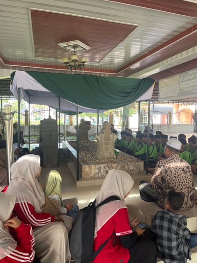 The image shows a group of people gathered at a tomb in Beuringen, Indonesia. The tomb, adorned with carved stone and covered in pebbles, is central to the scene. Several individuals, including women wearing hijabs and men in traditional attire, are seated or kneeling around the tomb. A group of young boys in green shirts and skullcaps are also present, seated attentively. Overhead, a green and white canopy provides shade, supported by poles. The surrounding environment includes a fence, greenery, and buildings with arched windows. A banner with text in Indonesian is visible on the right. The scene appears to be a religious gathering or a visit to a sacred site.