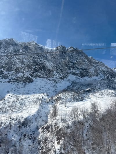 This media file presents a clear, high-angle view of a majestic winter mountain landscape in Alagna Valsesia, Italy. The scene is dominated by a vast, rugged mountain face, heavily covered in snow and ice, occupying the left and central parts of the frame. Dark rock outcrops are visible between patches of deep snow. A prominent cable, likely part of a cable car or ski lift system, runs vertically down the image from the upper left, appearing very close to the mountain.
The sky in the upper left is a brilliant, clear blue, indicating sunny weather. Reflections of internal structures or windows are visible on the glass through which the photo was taken, reinforcing the elevated vantage point, possibly from a cable car. To the right of the mountain, a snow-covered valley or lower slope descends, densely populated with numerous bare deciduous trees, characteristic of a high-altitude winter environment. These trees cast distinct shadows, suggesting bright sunlight during the morning or early afternoon.
No people or specific activities are visible within the frame; the image captures a serene natural vista. The overall impression is one of stark winter beauty, tranquility, and the grandeur of the snow-clad Alps. No text is visible in the image.