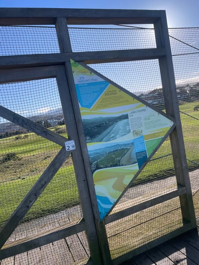 The image depicts a wooden viewing platform or railing on a clear, sunny day in Zarautz, Spain. A prominent diamond-shaped informational sign is affixed to the weathered wooden frame, which also incorporates a mesh netting. The sign presents text in multiple languages, including Basque, Spanish, English, and French, with titles such as "...AND A TRAPPED DUNE". It features aerial photographs of a coastline with a beach, urban development, and green areas, along with illustrative graphics in yellow, blue, and green. A small white label with the number "2" and a QR code is attached to a vertical wooden beam on the left. Faint graffiti is visible on a diagonal beam. The platform overlooks an expansive green, grassy landscape, possibly a golf course or natural area, extending towards distant hills and urban development on a hillside. The sky is clear blue, and strong sunlight from the right casts sharp shadows from the wooden structure.