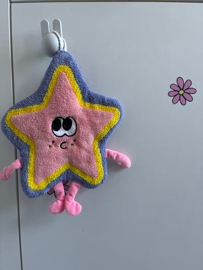 A pink, star-shaped sponge with a cartoon face is hanging from a grey bunny-shaped hook on a light grey, smooth surface, possibly a cabinet or locker door. The sponge has a purple border, a yellow inner border, and pink stuffing. It has black eyes with white pupils and eyelashes, rosy cheeks with three dots of green and red, and a small, curved mouth. Two pink arms extend from the sides of the star, and two pink legs dangle from the bottom. To the right of the sponge, on the same surface, is a sticker of a purple daisy with a yellow center. The overall scene suggests a child's room or bathroom in Haapsalu, Estonia.