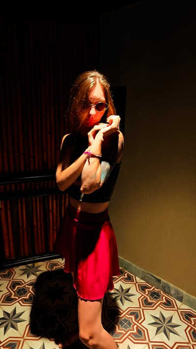 The image captures a single individual, appearing to be a young adult female, posing indoors under dramatic, directional lighting. She has long, light brown hair and wears dark-lensed, oval-shaped sunglasses, a dark crop top, and a bright red short skirt. Her forearms are crossed in front of her, with her hands resting near her shoulders. On her right wrist, a pink string bracelet and a multi-beaded bracelet are visible. Her left upper arm displays a multi-colored tattoo with intricate designs. The individual is barefoot. The background on the left consists of a dark, vertically slatted wooden or bamboo wall with a lower horizontal barrier, largely obscured by shadow. To the right, the background is a plain, dark-toned wall. The floor is covered with ornate geometric tiles featuring star and octagonal patterns in shades of brown, beige, off-white, and dark grey. A distinct, elongated shadow of the person is cast on the tiled floor, consistent with a strong light source positioned to her front-left. This scene is located in the city of Mu Cang Chai, Vietnam.