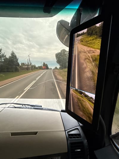 The image is taken from the interior of a vehicle, looking out through the windshield. The windshield is partially obstructed by the vehicle's interior, including a dashboard and a screen displaying a video feed. The screen shows a road with a white truck on it, and on either side of the road, there are green fields and trees. The sky is cloudy. The view through the windshield shows a similar road, with houses and trees on either side. The road has double white lines down the center. A red truck is visible in the distance on the road. The time of day appears to be daytime. The location is identified as Gulyayevka, Russia.