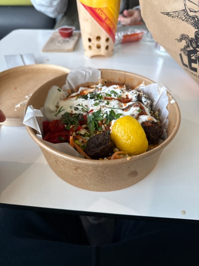 The image shows a take-away container, possibly from a restaurant chain, with food inside. The container is cylindrical and made of brown cardboard. Inside the container, there's food consisting of falafel balls, a half-lemon, a white creamy sauce drizzled with a reddish sauce, chopped herbs (probably parsley), and shredded vegetables, possibly carrots and pickled red peppers. The food is served on a white paper liner. The container is placed on a white table. Parts of paper packaging, including a yellow item with "LEON LEON" visible on it, are also on the table, indicating the restaurant or the brand. The background is slightly blurred. A black object is partially visible in the right side of the picture. The image seems to be taken indoors, possibly in a cafe or restaurant. Based on the Leon branding and the request for the location, it may be taken at the Leon restaurant location in Cannock Chase, United Kingdom.