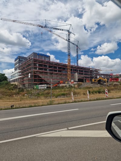 The image shows a construction site in Hechingen, Germany, under a partly cloudy sky. A large, multi-story building, partially covered in scaffolding, is the central focus. The building has "Bentley" branding visible on its facade. Multiple construction cranes are erected around the building, with one large crane extending high above the structure. In the foreground, a road with white lane markings runs across the bottom of the frame. To the right, a car's side mirror is partially visible, reflecting the road and some of the construction site. The surrounding area appears to be a grassy embankment leading up to the construction zone. A yellow excavator is visible on the construction site.