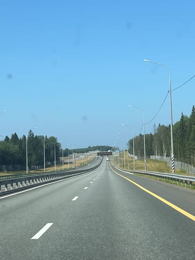 The image depicts a highway with a clear blue sky overhead. The road is paved and has white lane markings. There are streetlights lining the highway and a metal guardrail on the right side. Dense green trees are visible on both sides of the road, with a wooden fence in the foreground on the left. In the distance, a highway overpass can be seen. The scene appears to be a rural or semi-rural area, given the presence of trees and open space. The location is identified as Shchuch'e Ozero, Russia.