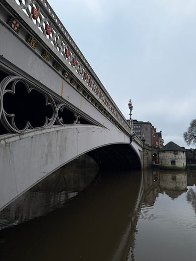 A low-angle shot of a bridge, emphasizing its architectural details. Ornate metalwork lines the bridge's side, featuring quatrefoil designs. A tower-like structure is at the top of the bridge. Its reflection is visible in the dark water beneath the arch. The sky is overcast, creating a gray backdrop. The composition is tilted, adding a dynamic perspective.
