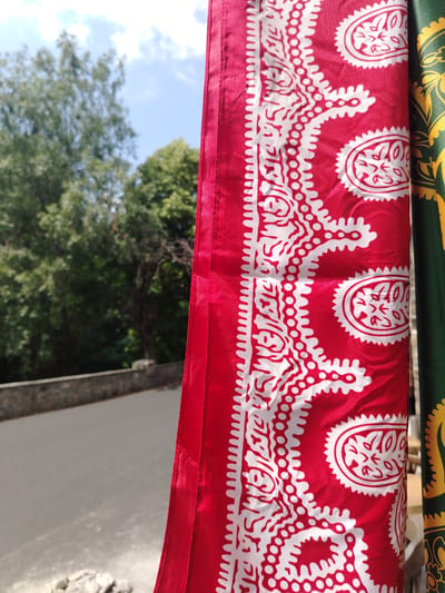 A close-up, horizontal shot focuses on a bright red fabric with a white, intricate paisley-like pattern, hanging vertically. To the right, a sliver of green fabric with a gold pattern is visible. The background is blurred, showing lush green trees, a bright blue sky with white clouds, and a paved road behind a low stone wall. The scene appears to be outdoors, possibly in a park or along a scenic route in the city of Shaki, Azerbaijan, given the distinctive traditional textile patterns often found in the region. No people are visible.