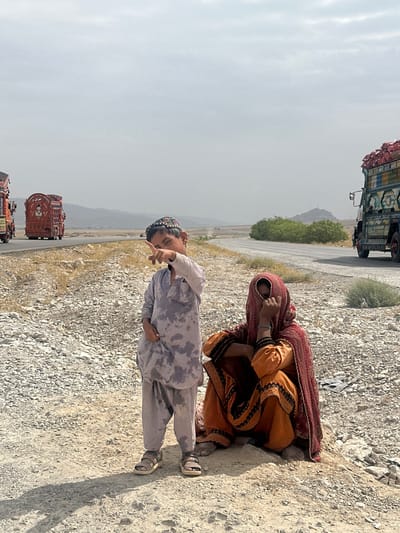 In Khanai Baba, Pakistan, a young boy stands on a rocky roadside, pointing into the distance with his right index finger. He wears a light-colored, patterned shalwar kameez and a traditional cap. Beside him, a woman is seated, covered by a maroon and orange hijab and clothing, with her hand near her face. The background shows a highway with several decorated trucks, mountains in the distance, and sparse vegetation. The sky is overcast.