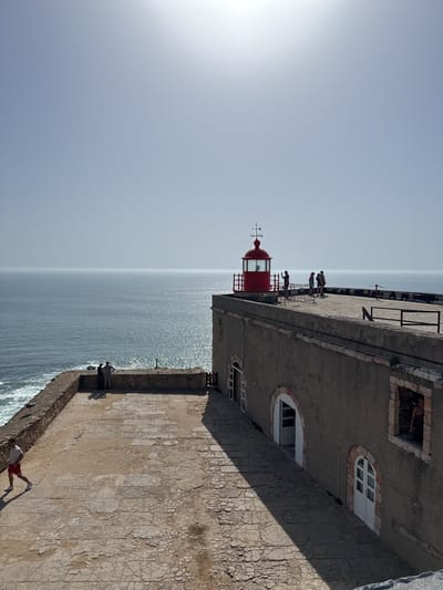 The image captures a scenic view of the lighthouse at Farol da Nazaré in Nazaré, Portugal. The scene includes the ocean stretching towards the horizon under a clear, bright sky. A stone structure dominates the lower portion of the frame, with a red lighthouse on top and several people standing near the edge of the structure. Other people are visible standing by the edge of the wall. A man in red shorts and a white shirt walks in the opposite direction. The structure has arched doorways and windows. The building casts a shadow on the textured, cracked stone surface in the foreground.