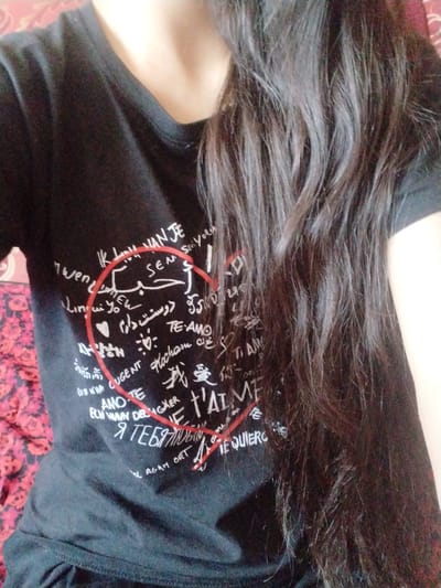 The image shows a close-up of a person wearing a black t-shirt with various phrases written in different languages, many of which appear to be "I love you" in various forms. A red heart is drawn over the text on the t-shirt. The person has long, dark hair that falls over their shoulder and partially obscures the t-shirt. The background is a blurry pattern of red and pink fabric. There is no indication of a specific location within the city of Социализм, Kyrgyzstan.