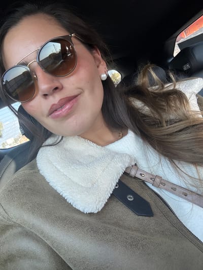 A woman wearing brown sunglasses and a single pearl earring is visible. She has medium-length brown hair. She's wearing an olive green jacket with a white, fluffy collar. The background is dark, possibly the interior of a car. A purse strap can be seen going over her shoulder. Her lips appear to have a glossy finish.