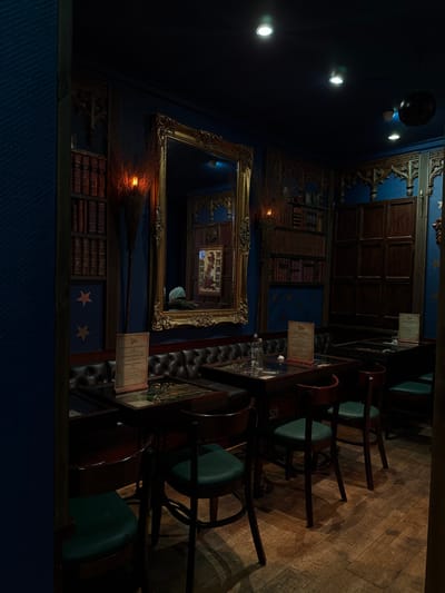 The image captures an interior view of a dimly lit, richly decorated room, likely a themed bar, cafe, or restaurant, presented in a portrait orientation but originally taken in landscape and rotated. The overall aesthetic suggests a magical or old-world library theme.
On the left side of the frame, a dark blue wall serves as a backdrop, adorned with several decorative elements. Faux bookshelves filled with numerous books are depicted on the wall, some appearing as wallpaper or painted murals, contributing to a library-like atmosphere. Large, ornate, gilded frames and mirrors are prominent features; one particularly large mirror prominently reflects another section of the room, creating an illusion of infinite depth with repeated reflections of frames and a small, framed picture within. Below this large mirror, a witch's broom or similar magical implement is affixed to the wall horizontally. Further down the wall, another section of faux bookshelves is visible, interspersed with two simple star decals. The wall also features dark wooden paneling at the top, adding to the classic, sophisticated feel. Small, warm-toned wall sconces or spotlights illuminate these decorative elements, casting a soft glow.
The right side of the image features a seating area. A long, dark wooden high-top table or bar counter extends across this section. Several dark wooden bar stools with round, dark green upholstered seats are neatly arranged along the table, suggesting a bar or casual dining setup. On the tabletop, multiple small, rectangular white cards in clear stands are visible, likely menus for the establishment. Adjacent to the high table, running parallel to it, is a dark, tufted leather or faux leather bench or booth seating, adding a touch of elegance and comfort. The floor in this area is made of polished wooden floorboards, contributing to the warm and inviting ambiance.
Overhead, on what appears to be a dark ceiling or upper wall, several small, bright spotlights are visible, providing additional illumination to the space. The lighting is generally subdued and atmospheric, creating a cozy and perhaps mysterious mood. A faint reflection of what might be a person or a light source is visible within one of the mirrors. The entire scene evokes a sense of stepping into a unique and carefully curated space.