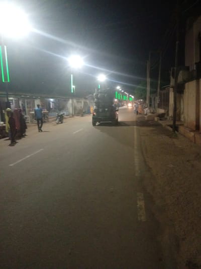 The image depicts a street scene at night in Tundun Wada West, Nigeria, illuminated by streetlights and the headlights of vehicles. A truck laden with what appears to be hay or straw is the central focus, driving down the asphalt road. On the left side of the road, several people are gathered near small shops or stalls. One man in a blue shirt and dark pants is walking away from the camera on the road. A motorcycle is parked near the shops. The right side of the road features buildings with a brick and stucco facade and utility poles with overhead wires. Green lights are visible on poles lining the street. The overall atmosphere is that of a bustling urban environment at night.
