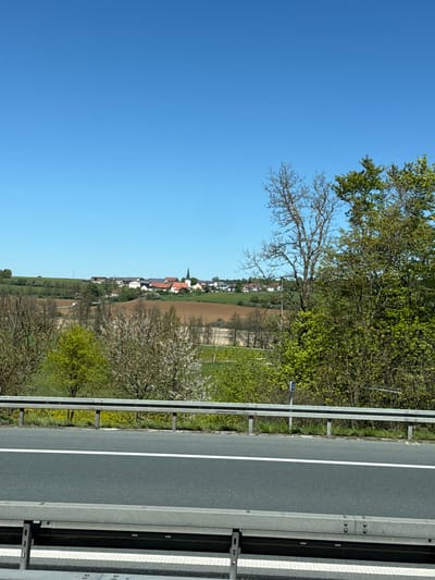The image depicts a roadside view in Velburg, Germany, featuring a highway with a metal guardrail along its edge. Beyond the guardrail, a landscape unfolds with trees in varying stages of foliage, a cultivated field, and a village in the distance. The village includes buildings with reddish roofs, one of which has a prominent steeple, possibly a church. The sky above is a clear, bright blue.