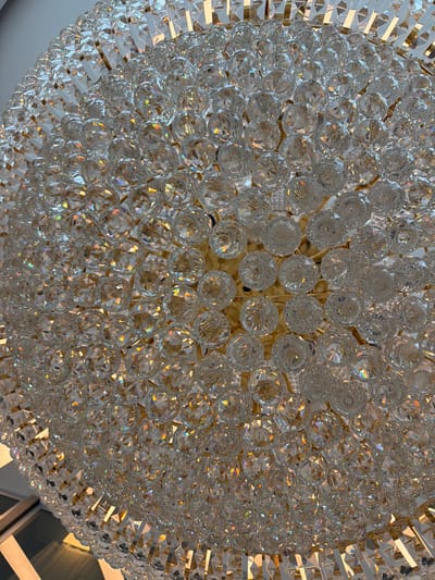 A close-up, overhead shot captures the intricate details of a large, spherical crystal chandelier. The chandelier is composed of numerous faceted crystal or glass balls, tightly packed together, creating a dazzling display of light and reflection. The crystals catch and refract light, producing a rainbow effect in some areas. A golden or brass colored frame is visible between the crystal elements, providing structure. The lighting suggests an indoor environment, possibly a grand hall or a luxurious room. The image does not contain any people or specific landmarks that would indicate its location in Ankara, Turkey.