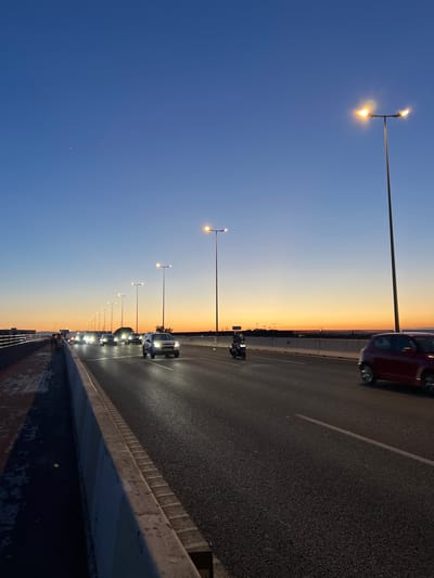 The image depicts a highway overpass at dusk, likely in Xirivella, Spain, characterized by a gradient of blue and orange hues in the sky. Several vehicles, including cars and a motorcycle, are in motion on the multi-lane road. Streetlights illuminate the highway, casting light on the wet asphalt surface. On the left side of the road, a pedestrian walkway is visible with a few individuals standing. The scene is framed by concrete barriers separating the walkway from the road and a distant cityscape with industrial structures.