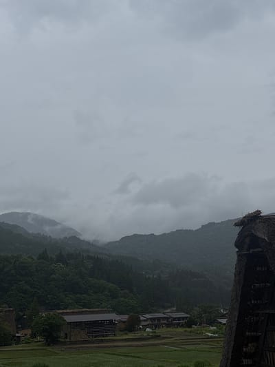 This image captures a wide view of a rural landscape in Shirakawa, Japan, under a heavily overcast sky. The upper half of the frame is dominated by thick, grey clouds, some exhibiting slightly lighter patches, suggesting an ongoing or recent period of rain. Below the sky, layers of densely forested mountains extend across the mid-ground, their peaks partially obscured by low-hanging mist and clouds, creating a subdued and atmospheric scene. The trees appear dark green due to the muted lighting.

In the lower half of the image, nestled at the base of the mountains, is a village characterized by traditional Japanese architecture and agricultural land. A significant portion of a large, dark, thatched-roof structure, consistent with the Gassho-zukuri style characteristic of Shirakawa-go, is prominent on the far right, partially cropped out of the frame. Its thick, straw roof and dark wooden framework are visible. Towards the center-left, several other buildings are interspersed among trees and green fields. One notable structure features a dark roof and an open side with horizontal wooden slats, possibly for drying or storage. The foreground consists of vibrant green rice paddies, segmented by narrow water channels, indicating active cultivation. No people or animals are visible in the scene, which conveys a sense of quiet rural life.