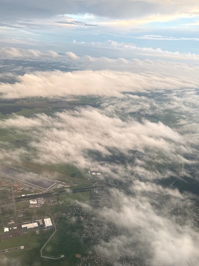 This is an aerial, high-angle, medium shot of clouds partially obscuring the landscape of Aramil, Russia. The clouds are predominantly white with hints of pink and yellow from the sky. The land below is a mix of green fields, dark forests, and urban areas. We can identify a long, striped runway, likely an airport or airfield, and several rectangular buildings, possibly industrial or commercial structures. Smaller residential areas with houses are also visible. The clouds create a dynamic, layered effect, with some appearing wispy and stretched, while others are more voluminous. The lighting suggests it may be sunrise or sunset due to the warm tones in the sky and the soft illumination of the clouds.