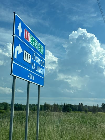 A blue road sign with white text and symbols is pictured against a bright blue sky with large, white cumulus clouds. The sign, mounted on three metal poles, directs traffic. The top section shows an upward arrow and the route numbers "11" (in red and white), "E 265" (in green and white), and "E 67" (in green and white). The middle section indicates a right turn with the route number "117" (in white on a black background) and the destination "JÕGISOO VALINGU". The bottom section indicates "400m". The sign is situated in a grassy field with tall, green grass. In the background, a line of trees is visible, with a power line running across the horizon. The overall scene suggests a rural or semi-rural setting in Tutermaa, Estonia. There are no people visible in the image.