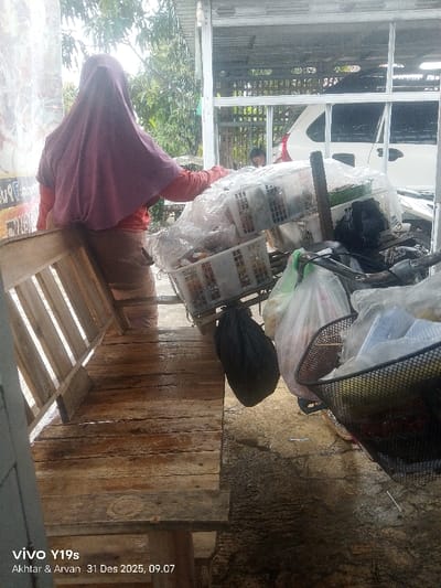 An outdoor scene in Jagapura Kidul, Indonesia, shows a person facing away from the camera, wearing a pink hijab over an orange long-sleeved shirt and light-colored pants. This individual is leaning against a weathered wooden bench on the left and appears to be attending to a heavily loaded pushcart positioned in the center. The pushcart contains multiple white plastic crates filled with various items, largely covered in clear plastic sheeting. Several additional transparent plastic bags and a black plastic bag are hanging from the cart's frame, which also features a black wire mesh basket overflowing with contents on the right side. The handlebars of the cart are visible. In the background, a white vehicle (possibly an SUV or minivan) is parked or partially visible behind a metal-framed structure. A second individual, appearing to be a male child or young person in a dark shirt, is partially visible in the distant background. The ground is an uneven, damp surface, and green foliage is visible beyond the structure. The lighting suggests daytime conditions.