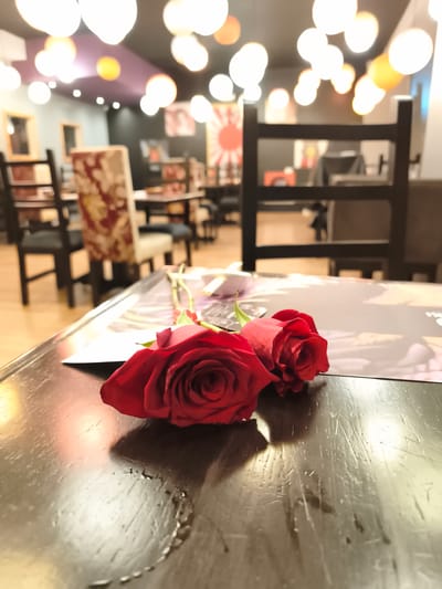 The image shows two red roses laying on a dark wood table in a restaurant. In the blurred background, there are tables and chairs, and the ceiling is decorated with round, glowing lights. A Japanese-style rising sun flag is visible on the wall. This is likely a restaurant in Quito, Ecuador.