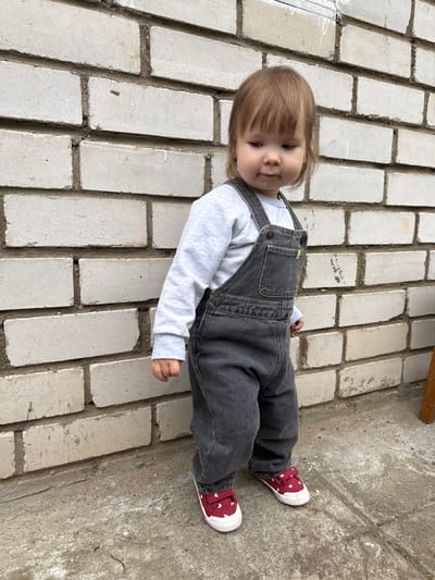 The image shows a young child standing in front of a white brick wall. The child is wearing a light gray long-sleeved shirt and dark gray overalls. The child also has on red canvas shoes with white soles and white heart patterns. The child's hair is light brown and styled with bangs. The child is looking down and to the right. The brick wall appears to be part of an outdoor structure. The ground beneath the child is a concrete surface. The image is taken in Vyshegor, Russia.