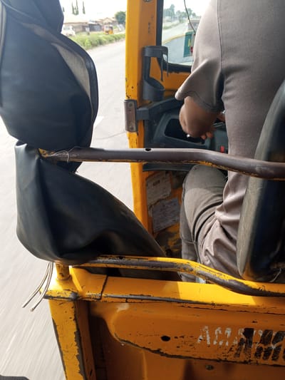 The image shows the interior of a yellow auto-rickshaw, likely a commercial vehicle, with a driver's hand on the steering wheel. The driver is wearing a grey t-shirt and grey trousers. The auto-rickshaw appears to be in motion on a paved road with some vegetation and buildings visible in the background. The interior is utilitarian, with black vinyl seats and exposed metal framing. Some of the paint on the exterior of the auto-rickshaw is chipped and worn, suggesting it is an older vehicle. The text "AGAMAMA" is visible in black paint on the yellow body of the auto-rickshaw. Based on the text and the general appearance of the vehicle and surroundings, the location is identified as Ashata, Nigeria.
