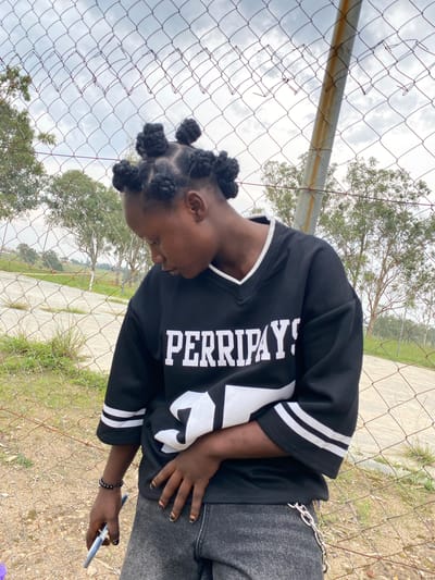 In Zawan, Nigeria, a young Black female is captured in a medium shot, leaning against a chain-link fence. Her head is tilted downwards and to her left, and her dark hair is styled in multiple rounded Bantu knots. She is wearing a black, V-neck, short-sleeved jersey with white trim and bold white lettering across the chest that reads "PERRIPAY!". The jersey also features white stripes on the sleeves. Her lower half is clad in dark wash, distressed denim jeans with a silver chain attached to the front belt loop. She is holding a light blue object in her right hand, which is resting on her hip. Her left hand is also on her hip, near her jeans. A black beaded bracelet adorns her left wrist. The background reveals a cloudy sky, trees, and a paved area, possibly a sports court, visible through the diamond-shaped openings of the fence. The ground in the foreground consists of gravel and dry grass.