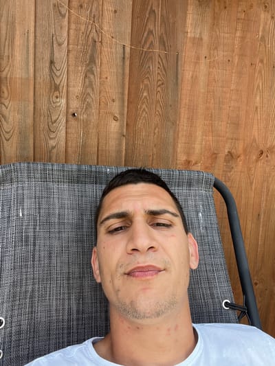 A man lies on a grey lounger in an outdoor setting. The lounger has a black metal frame. He wears a white shirt. The background is a wooden wall. No location information is available.