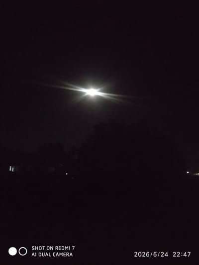 The image depicts a night scene in Khalkabad, Uzbekistan. The sky is dark, with a bright light source at the top center, likely a street light or the moon, casting a starburst effect. In the foreground, silhouetted trees and some indistinct structures are visible, suggesting an outdoor or semi-rural setting. There are faint lights from what appear to be buildings or distant lights.
The time of day is clearly indicated by the timestamp in the bottom right corner: 2026/6/24 22:47, confirming it is nighttime. There are no people visible in the image. The overall mood is dark and quiet, with the bright light providing the only significant illumination.
Text overlay at the bottom left reads "SHOT ON REDMI 7 AI DUAL CAMERA", indicating the device used to capture the photograph.