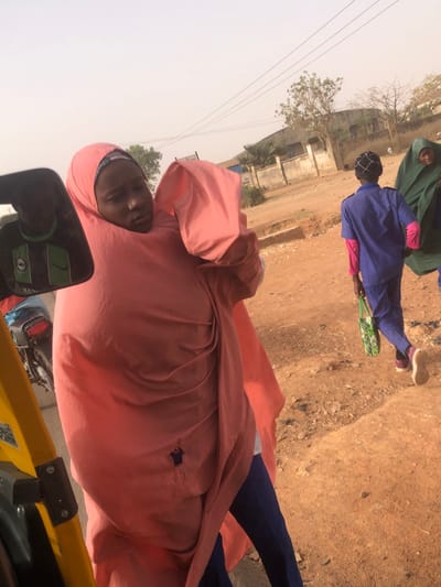 A person wearing a peach-colored hooded garment is visible, partially obscured by a vehicle mirror. Other people in blue and green garments walk on a dirt road alongside buildings and a tree. The sky appears hazy.