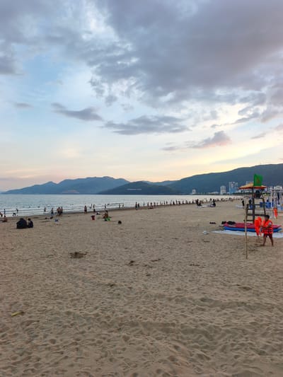 The image depicts a beach scene in the city of An Nhơn, Vietnam. The foreground is dominated by a wide expanse of light brown sand, textured with numerous footprints and small mounds. In the middle ground, a body of water, likely the sea, stretches out, with a gentle surf breaking near the shore. Numerous people are present on the beach and in the water. Some are walking along the shoreline, while others are wading or swimming. A lifeguard stand is visible on the right side of the image, equipped with a green flag and a life vest. Two red life vests are also seen on the sand near the lifeguard stand, along with two inflatable rafts. A young boy wearing red shorts and an orange life vest is standing near the lifeguard stand. In the background, a long line of hills or mountains forms the horizon, with some buildings and vegetation visible along their slopes. The sky above is partly cloudy, with shades of blue and grey, and hints of pastel colors suggesting either dawn or dusk. The overall atmosphere is one of a recreational beach setting.