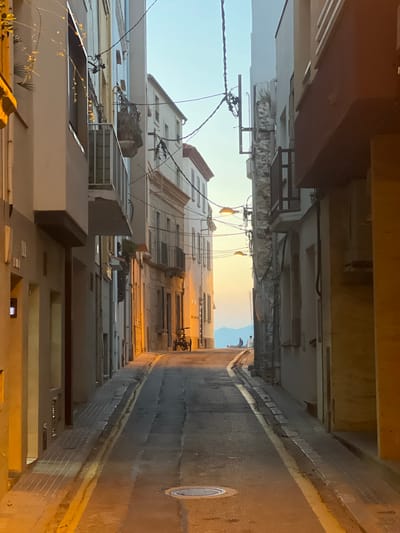 The image depicts a narrow street in L'Escala, Spain, at what appears to be either dawn or dusk, given the warm lighting and soft sky. The street slopes gently uphill and is flanked by multi-story buildings with balconies, some displaying potted plants. The architecture suggests a Mediterranean style.
In the foreground, the asphalt road is marked with yellow lines. A circular manhole cover is visible in the lower center. To the left, a textured beige building features a dark rectangular window or panel. Further up the street, on the right side, a bicycle is parked near the curb.
As the street recedes into the distance, the sky transitions from a pale blue at the top to a warm orange and yellow hue near the horizon, indicating the presence of the sun. In the far distance, silhouetted against the bright sky, are two figures standing near a body of water, and the faint outline of mountains. Several streetlights are mounted on poles and wires that crisscross overhead, illuminating parts of the street with a warm glow. The overall impression is a quiet, potentially residential, street in a coastal Spanish town.
