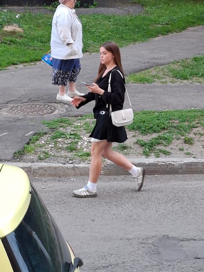 The image shows two people on a street in Bel'kovo, Russia. In the foreground, a young woman is walking from left to right, looking at her phone. She is wearing a black long-sleeved shirt, a black pleated skirt, white socks, and patterned sneakers. A light-colored crossbody bag is slung over her right shoulder. Behind her, an older woman is standing, facing to the right. She is wearing a white jacket over a dark top and blue patterned pants, with white shoes. A blue bag is hanging from her left hand. The street is paved with asphalt, and there is a patch of grass and gravel to the right of the paved area. A yellow car is partially visible in the bottom left corner of the frame. A manhole cover is visible on the pavement in the middle left of the image.