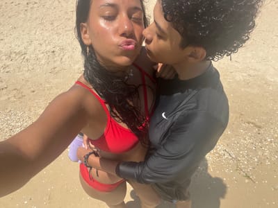 On a sandy beach in Unknown, Venezuela, a dark-skinned male with curly hair, wearing a black long-sleeved shirt and shorts, lies near a dark-skinned female with wet, dark hair, wearing a red bikini. They are posed close, with the woman's lips pursed and eyes closed as if preparing for a kiss. The woman is holding a purple bottle in her left hand. The background is beige sand with scattered debris. The photo appears to be a selfie taken from an overhead angle.