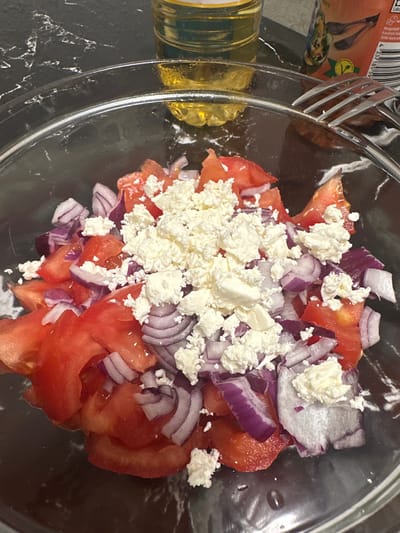 A close-up, high-angle shot captures a vibrant salad in a clear glass bowl, likely in an indoor kitchen setting in Fulda, Germany. The salad is composed of diced red tomatoes, chopped red onions, and a generous topping of crumbled white feta cheese.

To the left of the bowl, a metal fork rests partially against the bowl's rim and an orange-red product label. Further left, a clear bottle containing a yellow liquid, probably olive oil, is visible. The surface beneath the bowl and bottle is a dark, possibly black or dark grey, countertop with a subtly marbled or speckled pattern.

The scene depicts a salad in the process of being assembled or prepared, suggesting a fresh, simple meal. Visible text on the orange-red label includes "Hergestellt f[ür]" (Manufactured for) and "Friedrich F[elix]", along with a barcode starting "4 337185 59" and a small green logo that appears to be an eco or organic certification mark, consistent with German product packaging. The focus is on the fresh, colorful ingredients, highlighting the visual appeal of the dish.