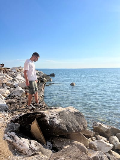 A young man with short dark hair and sunglasses stands on a rocky shoreline in Potos, Greece. He is wearing a white t-shirt and grey cargo shorts. He is looking down at the water, which is calm and blue. The shore is made up of large white and grey rocks, with some smaller pebbles and sand. The sky is a clear, bright blue.
