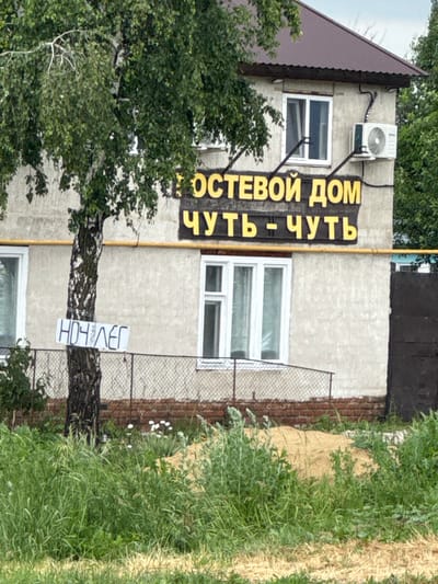 This is a medium shot, slightly high-angle, eye-level photograph of a building with a sign that reads "ПОСТЕВОЙ ДОМ ЧУТЬ-ЧУТЬ" in yellow Cyrillic letters against a dark background, positioned above another line of text. The building appears to be a single-story structure with a dark brown pitched roof. There are several windows visible on the facade, some with white frames. An air conditioning unit is mounted on the exterior wall next to a window. A birch tree with a thin trunk and green foliage stands to the left of the building, partially obscuring it. A white sign with black Cyrillic lettering, reading "НОЧЛЕГ," is attached to the birch tree. A yellow pipe runs horizontally across the building's facade. A chain-link fence with a red brick foundation is visible in front of the building. The foreground is filled with green grass and vegetation. The sky is overcast and appears to be daytime. The scene is located in the Voskresensky District, Russia.