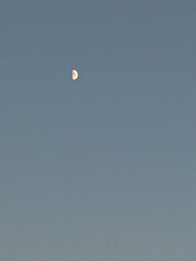 The image shows a clear sky with a waning crescent moon visible in the upper left quadrant. The sky is a uniform light blue, with no clouds or other celestial bodies apparent. There are no visible landmarks, structures, or people in the image. The scene is characteristic of an outdoor daytime or early evening sky.