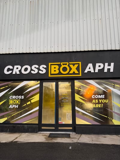 The image shows the storefront of "Cross Box Aph," a fitness center. A black sign displays the name vertically with "Cross" and "APH" in white, and "BOX" in a yellow square. The entrance has two glass doors with a graphic design of white, yellow, and purple stripes. The words "Come As You Are!" are displayed on the upper part of the window. Opening hours for Monday-Friday (06:30-20:30) and Saturday (10:00-20:00) are listed on the glass.